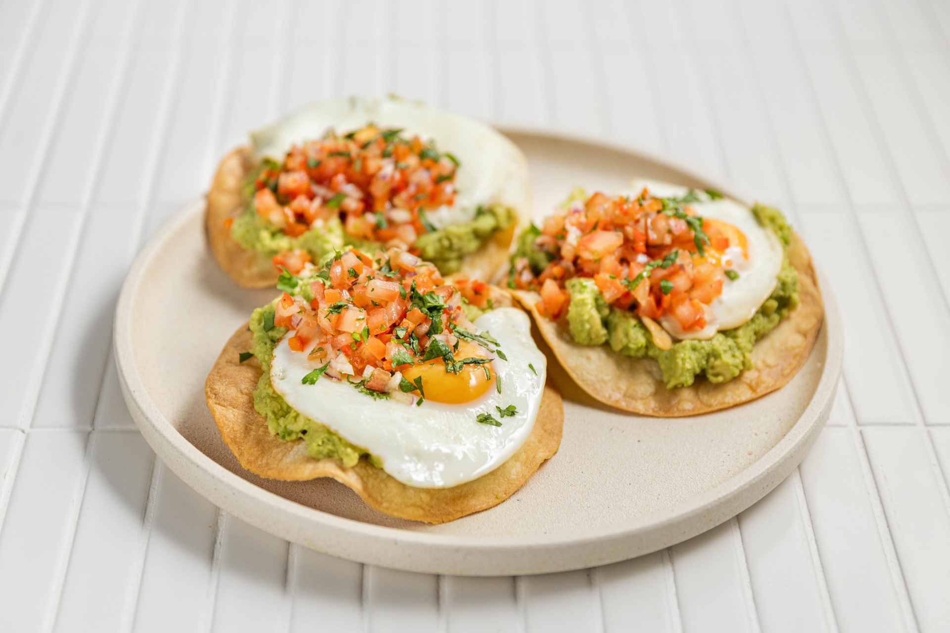 A plate of gluten free breakfast tostadas, featuring fried eggs, avocado, and chopped tomato with seasoning.